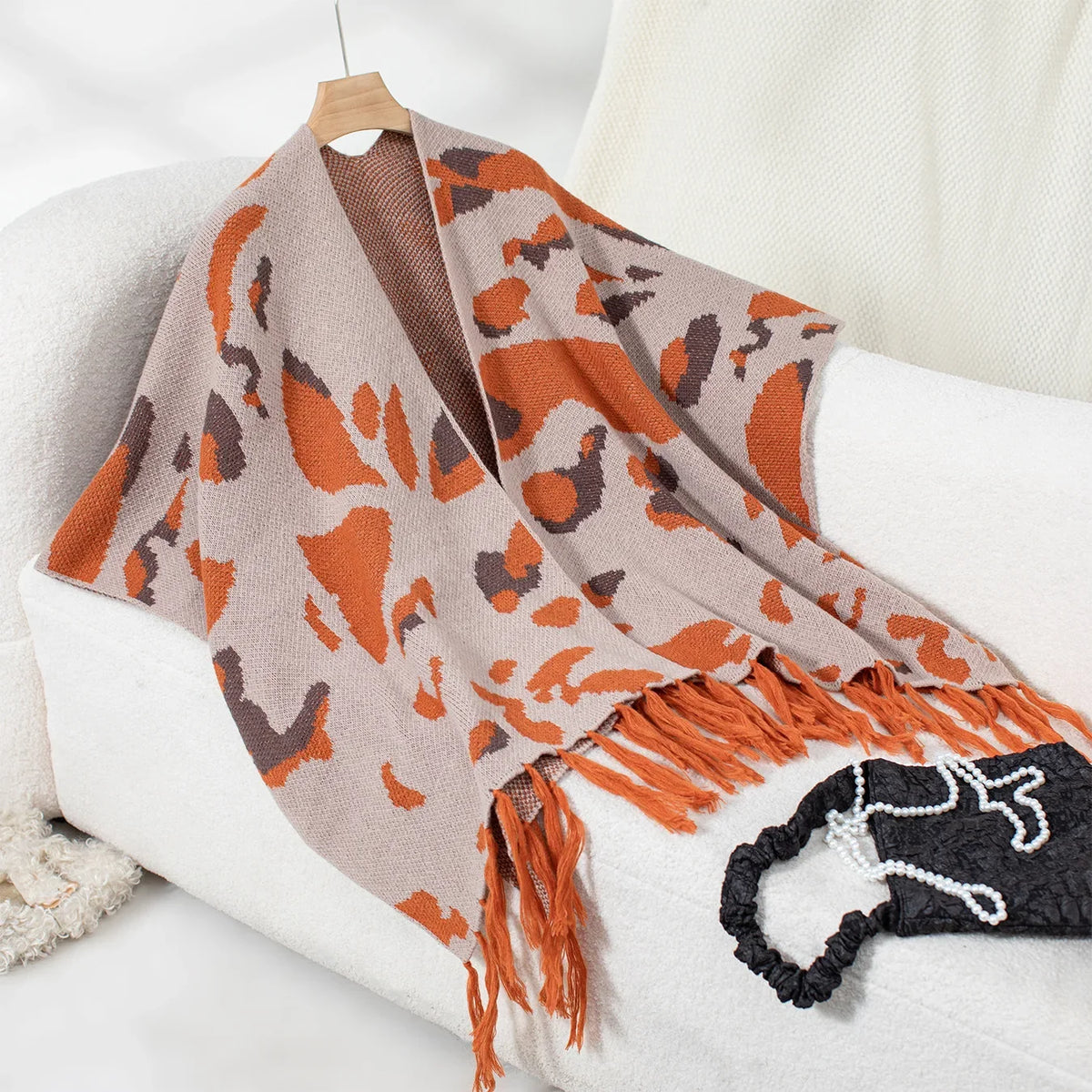 Cape poncho femme léopard orange et taupe avec franges, présentée sur cintre et chaise blanche