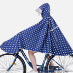 Femme à vélo portant un poncho de pluie bleu marine à pois blancs avec capuche et visière