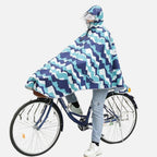Femme à vélo portant un poncho pluie bleu turquoise à motifs géométriques avec capuche et visière
