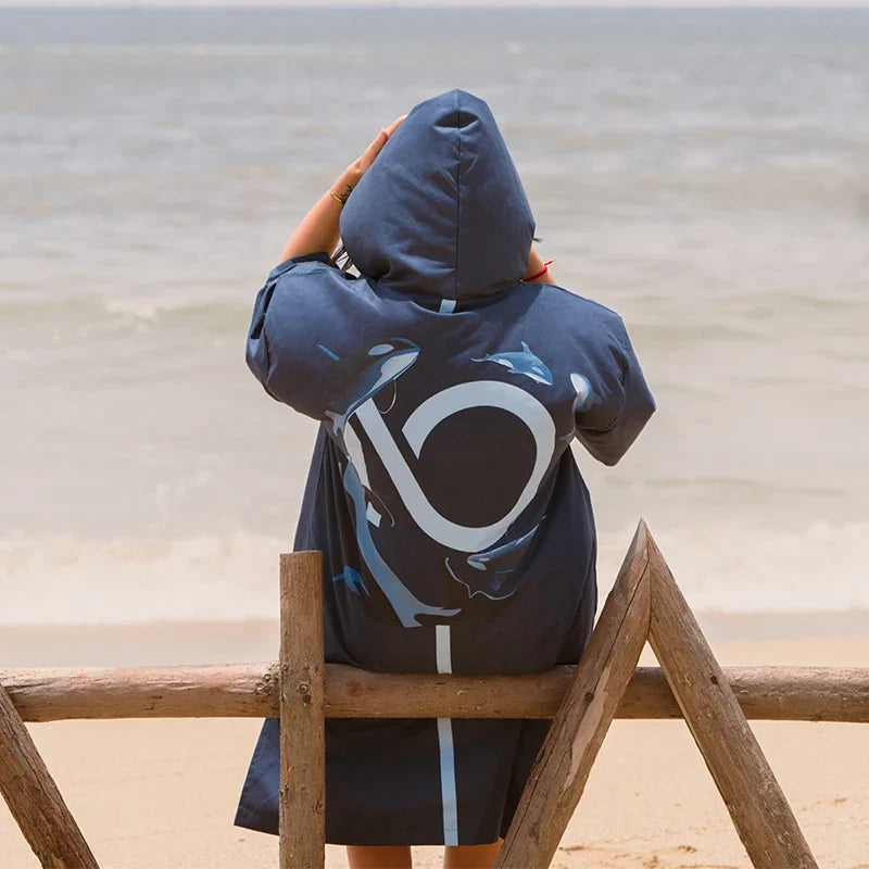 Femme de dos portant poncho surf bleu marine à capuche et poches, assise sur plage